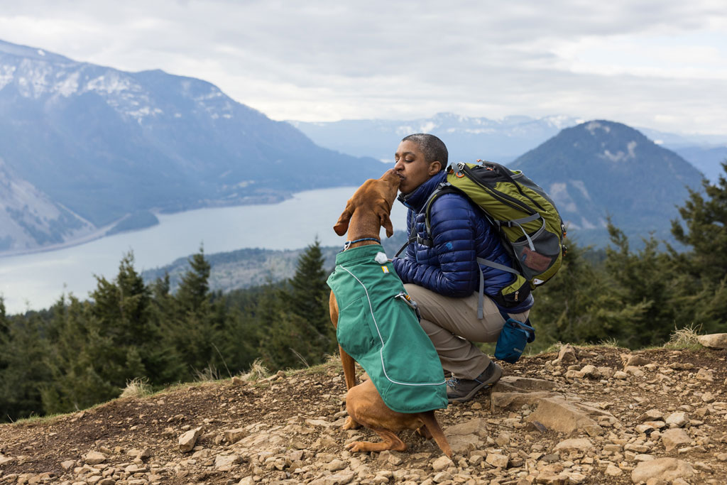 Kronda squats at the top of a hiking trail. Her Vizsla is sitting next to her giving her a kiss as they enjoy the view. 